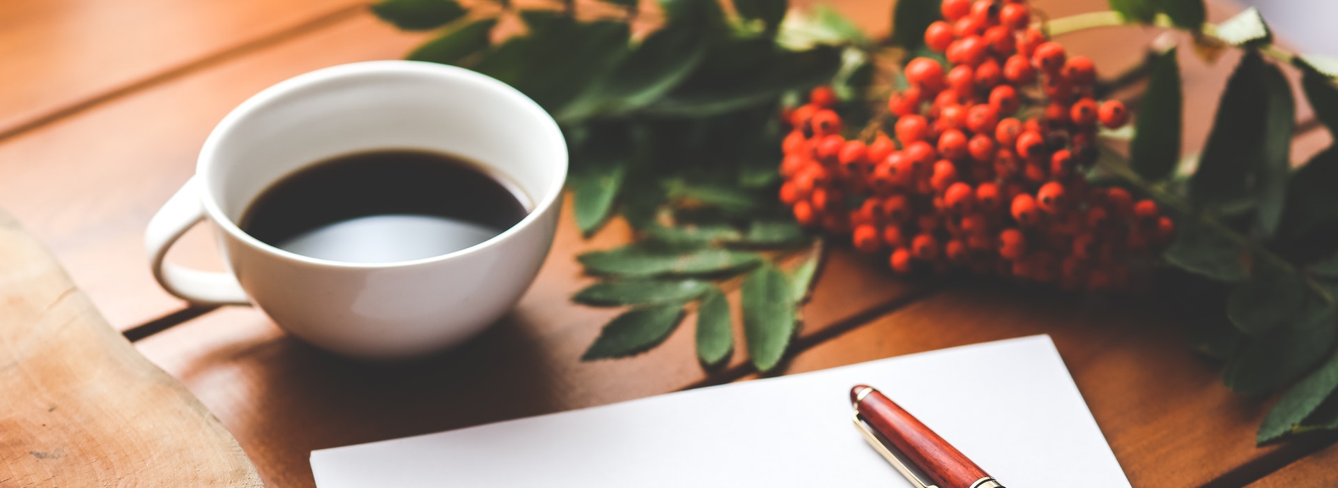 A white cup of coffee, a sheet of blank paper and some autumn foliage lay across a wooden desk