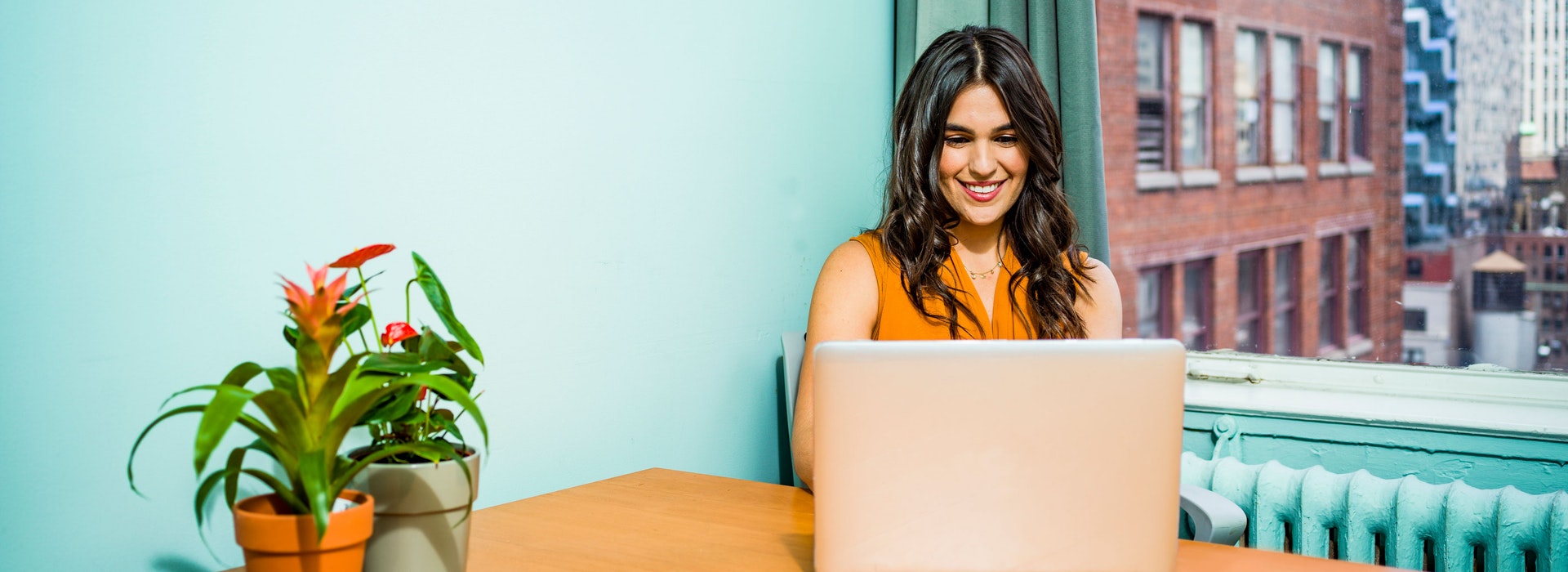 Femme aux longs cheveux foncés assise devant un mur turquoise qui sourit en regardant son ordinateur portable