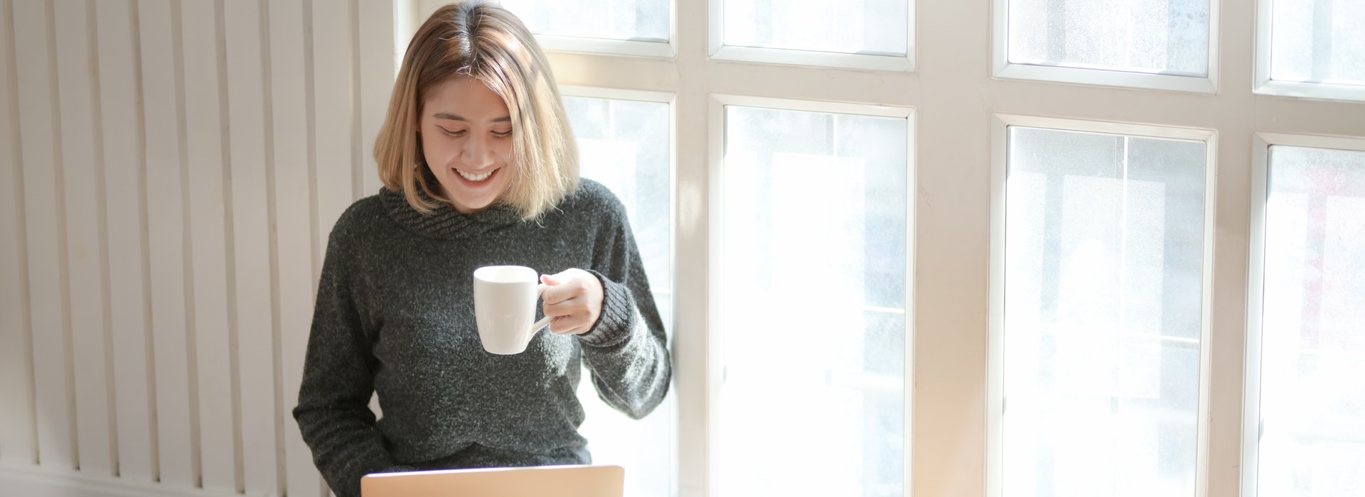 Une femme assise à une fenêtre boit du café et tape sur un ordinateur portable
